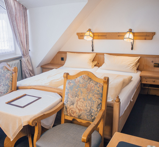 Helles Doppelzimmer mit elegantem Holzbett, zwei historischen Sesseln am Fußende und viel Tageslicht – charmantes Hotelambiente.